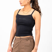 A woman wearing a black square-neck tank top with thin straps and a small white script logo on the chest, paired with brown pants, photographed against a plain white studio background.